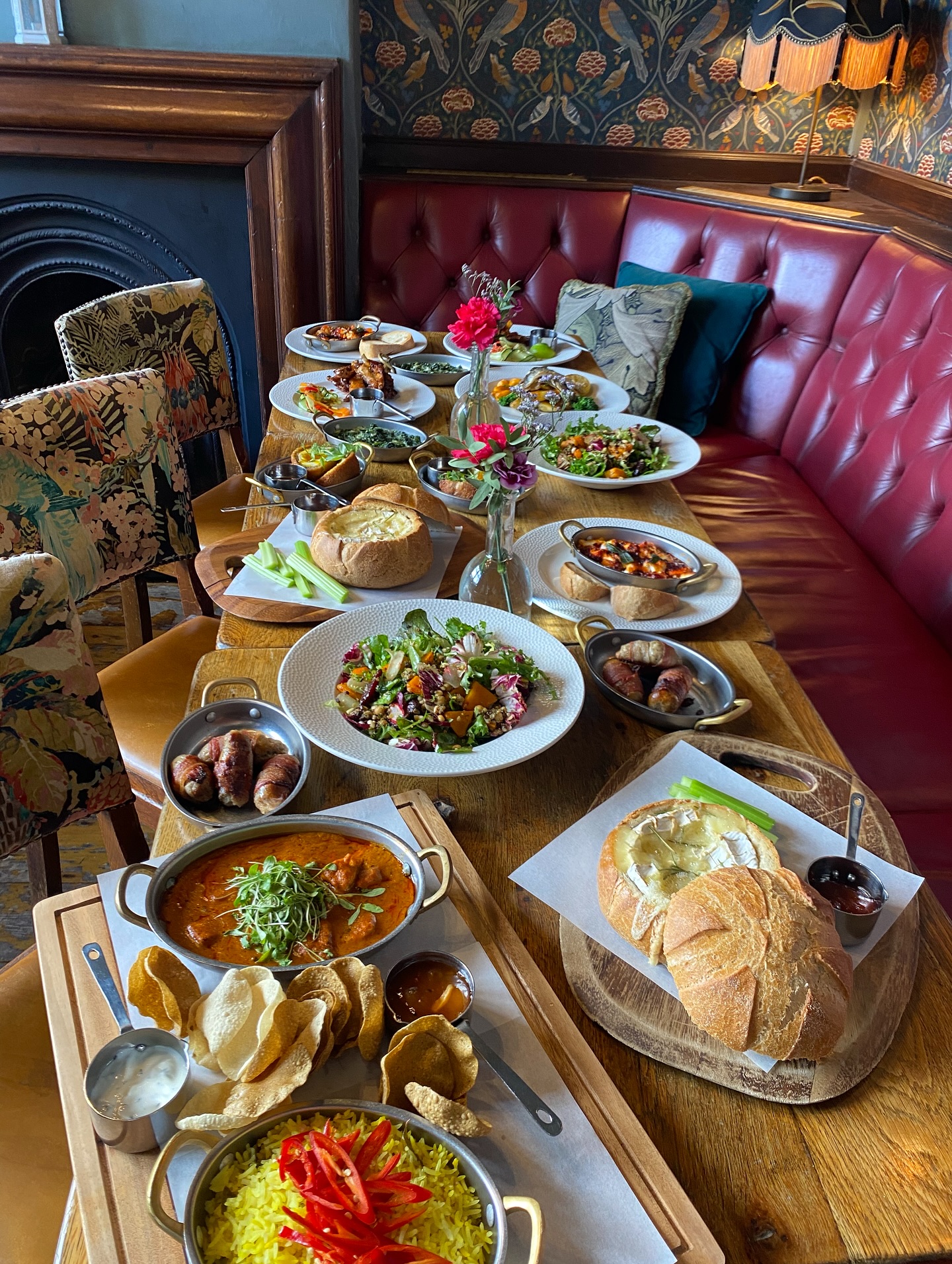 A large spread of dishes arranged across a long wooden table, including salads, curries, bread bowls, small plates and appetisers, set in a booth-lined pub interior.