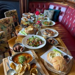 A large spread of dishes arranged across a long wooden table, including salads, curries, bread bowls, small plates and appetisers, set in a booth-lined pub interior.