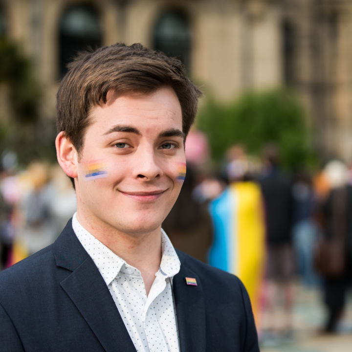 A person wearing a dark blazer over a white patterned shirt is standing outdoors in front of a blurred crowd and historic building. A small rectangular pin with horizontal stripes is attached to the blazer lapel. The background includes greenery and people, some holding colorful flags.