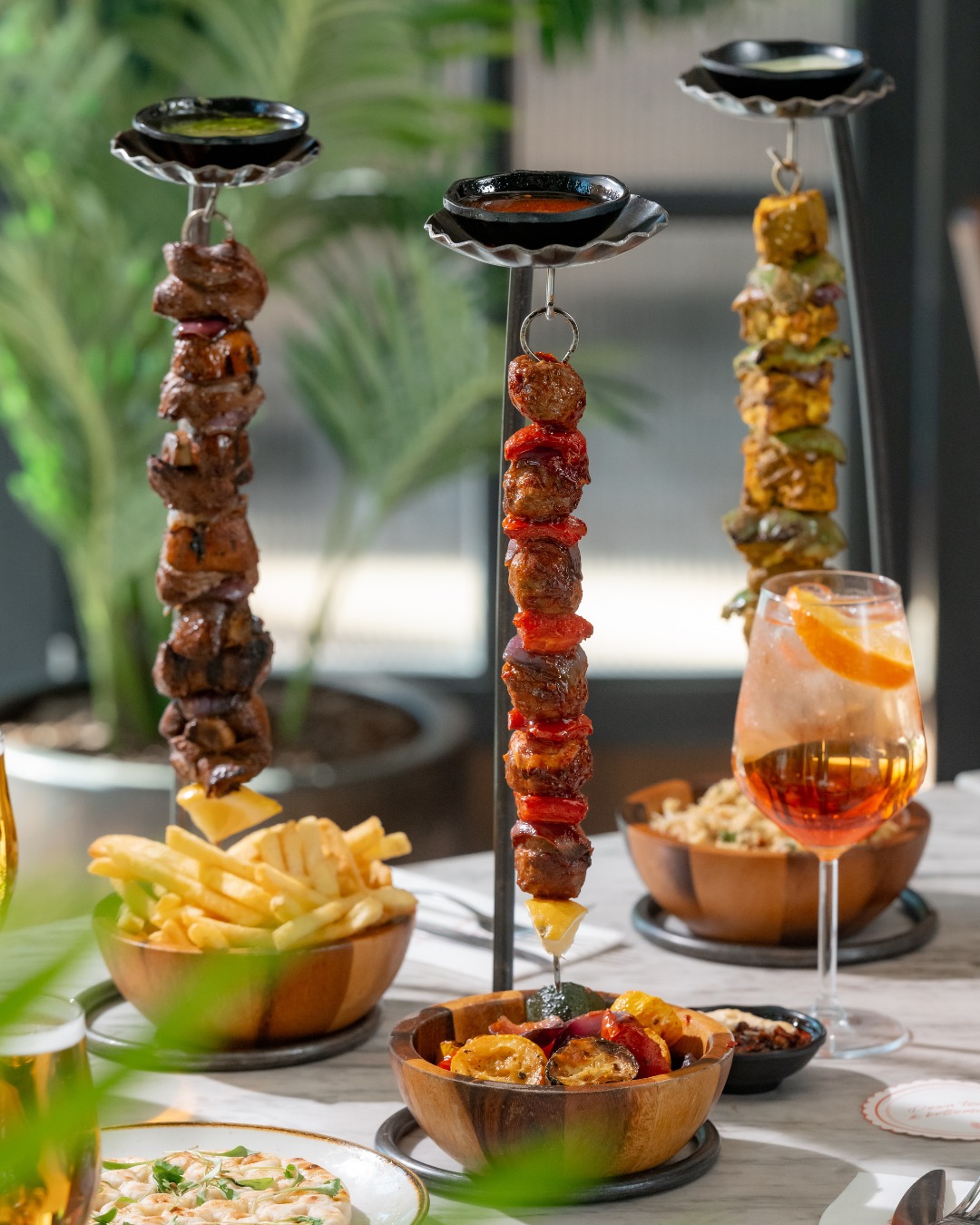A close-up of skewered meats and vegetables suspended on tall metal stands, served alongside bowls of fries and other dishes, with a cocktail on the table.