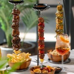 A close-up of skewered meats and vegetables suspended on tall metal stands, served alongside bowls of fries and other dishes, with a cocktail on the table.