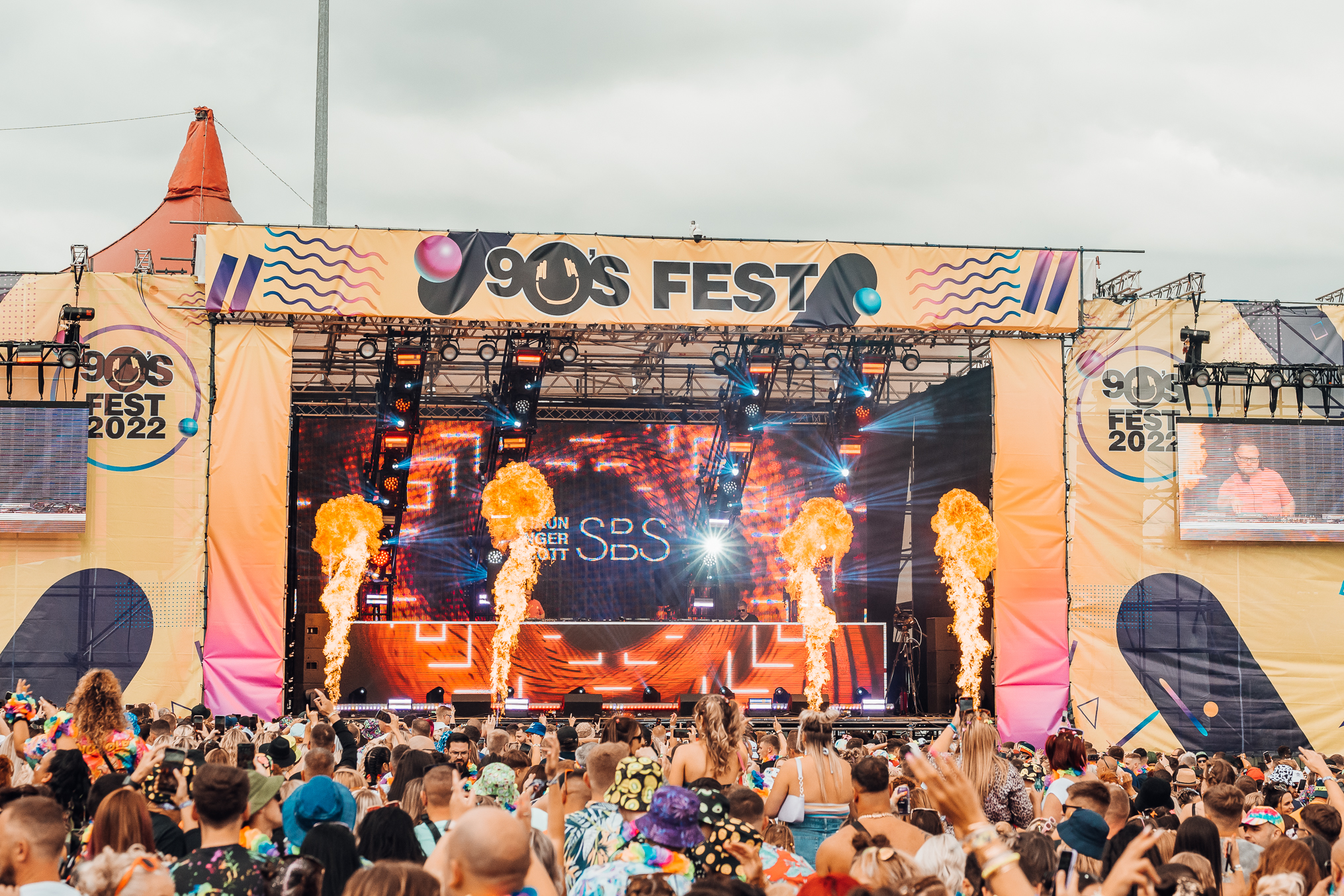 Looking across crowd towards 90s Fest Stage in the sunshine with pyrotechnics sending flames into the sky 