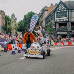 A soapbox cart designed to look like a cow, complete with black-and-white spots and a cow head on the front, is being pushed by a person wearing an orange jacket. Another participant in a cow costume sits inside the cart, raising one arm. The cart has a yellow plate with the text “ADAM 01.” The race takes place on a city street lined with safety barriers, purple-and-white cones, and directional signs, with a large crowd of spectators behind metal railings. 