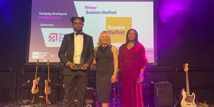 A group of people dressed in formal attire posing together at an indoor event with blue and purple stage lighting. Behind them is a stage with musical instruments and a large screen displaying “Sheffield Business Awards 2023” along with sponsor logos. One person in the center is holding an award, and another on the right is holding a framed certificate. The setting suggests a celebratory awards ceremony.