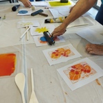 A table covered with a white cloth during a printmaking workshop. Several hands work with ink rollers, brayers and carved printing blocks. Brightly coloured ink—yellow, blue, red and orange—is spread on printing plates, and multiple printed artworks featuring bold floral designs are laid out across the table.