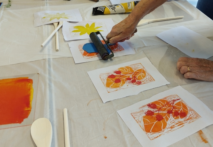 A table covered with a white cloth during a printmaking workshop. Several hands work with ink rollers, brayers and carved printing blocks. Brightly coloured ink—yellow, blue, red and orange—is spread on printing plates, and multiple printed artworks featuring bold floral designs are laid out across the table.