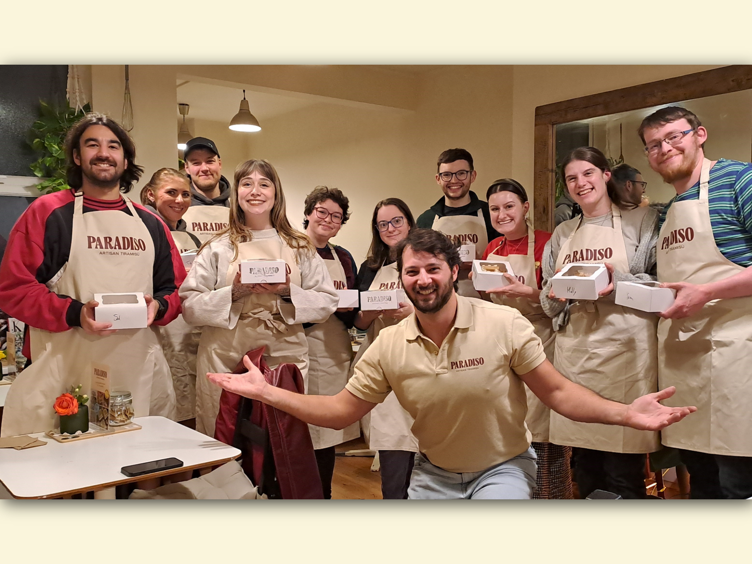Group of people standing indoors wearing beige aprons with “PARADISO” printed on them, holding white pastry boxes. A person in front kneels with arms open, wearing a matching beige shirt with the same logo. The setting includes wooden tables, chairs, a small flower arrangement, and warm lighting, suggesting a baking or cooking class environment.
