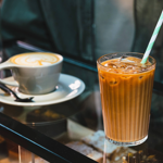 A close-up of two drinks on a glass counter in a coffee shop: a grey ceramic cup of latte with foam art on a saucer and spoon, and a tall glass of iced coffee with a green-and-white striped paper straw. Behind the drinks, part of a person’s jacket is visible, and jars of biscuits can be seen in the background.
