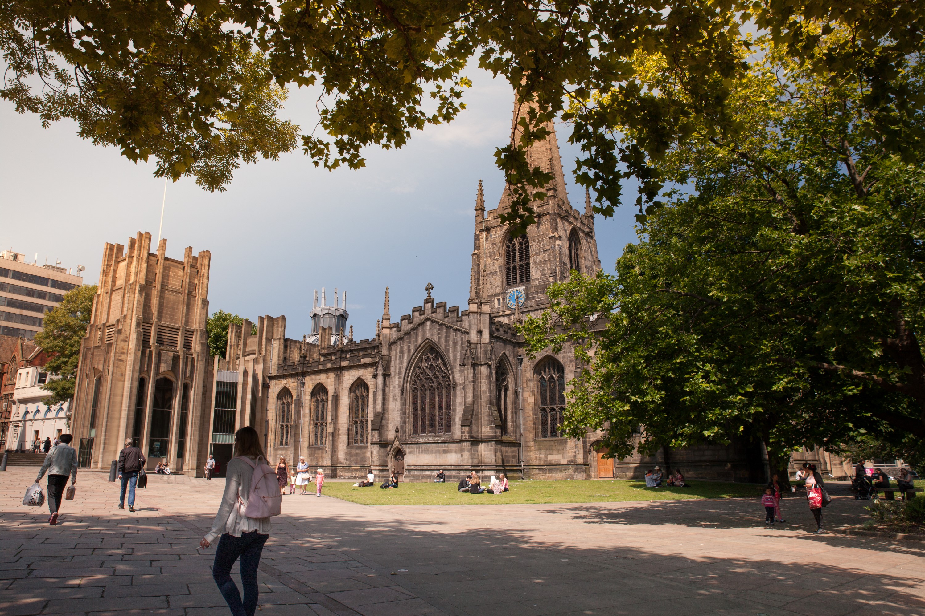 Sheffield Cathedral viewed from a shaded square, with large trees framing the scene. The historic stone building features a tall spire, ornate windows, and Gothic architecture. People are walking and sitting on benches in the sunlit open space in front of the cathedral.