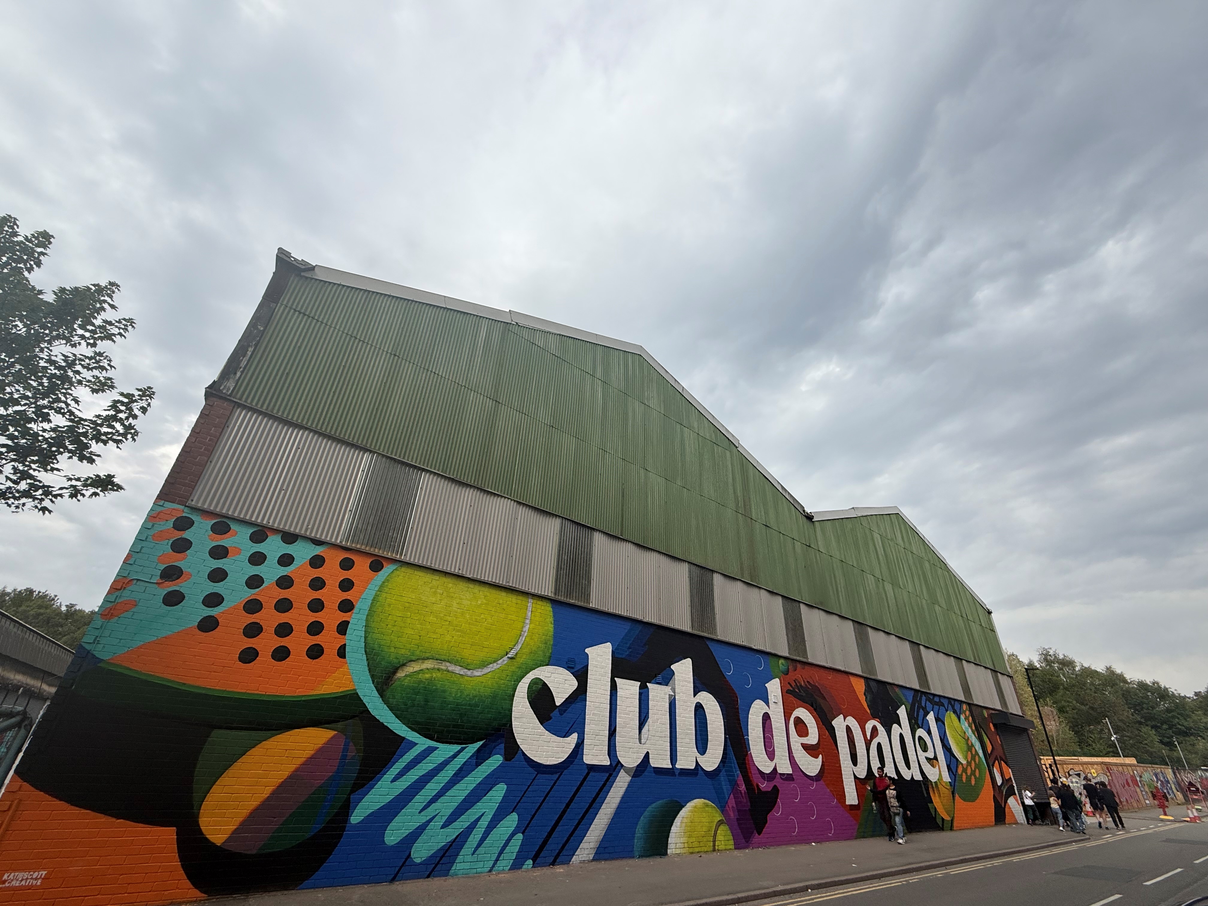 A large industrial-style building with green corrugated metal roofing and walls, featuring a vibrant, colorful mural along the lower section. The mural includes abstract shapes, bright colors, and large text reading “club de padel” in white letters. Several people are walking along the street in front of the building under a cloudy sky.
