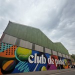 A large industrial-style building with green corrugated metal roofing and walls, featuring a vibrant, colorful mural along the lower section. The mural includes abstract shapes, bright colors, and large text reading “club de padel” in white letters. Several people are walking along the street in front of the building under a cloudy sky.