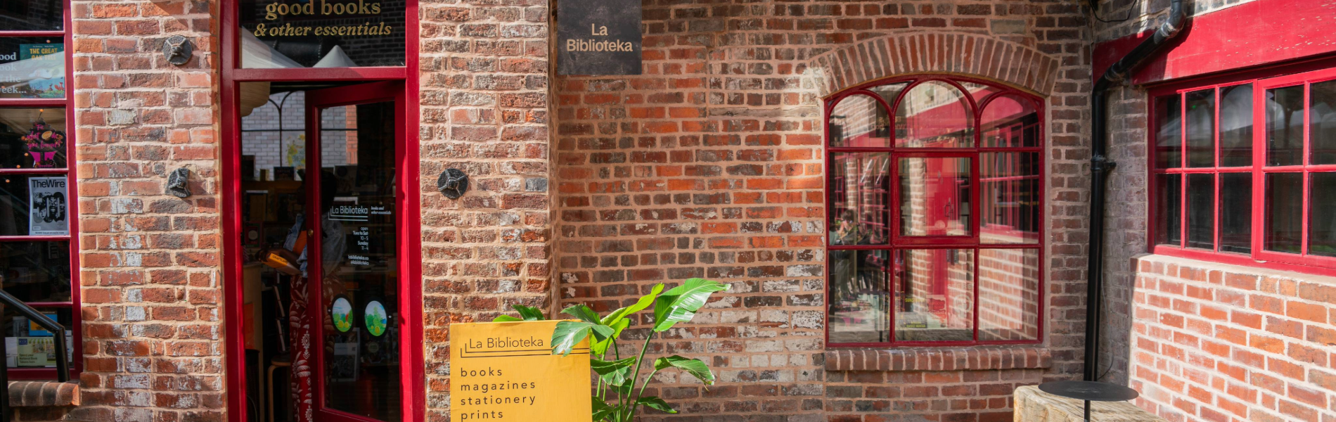 Exterior of a brick building with a red-framed door and windows. The entrance has a sign above reading “good books & other essentials,” and a hanging sign says “La Biblioteka.” A bright yellow board outside lists items such as books, magazines, stationery, prints, and gifts. A green potted plant sits beside the board on a cobbled walkway, creating a welcoming and vibrant storefront.