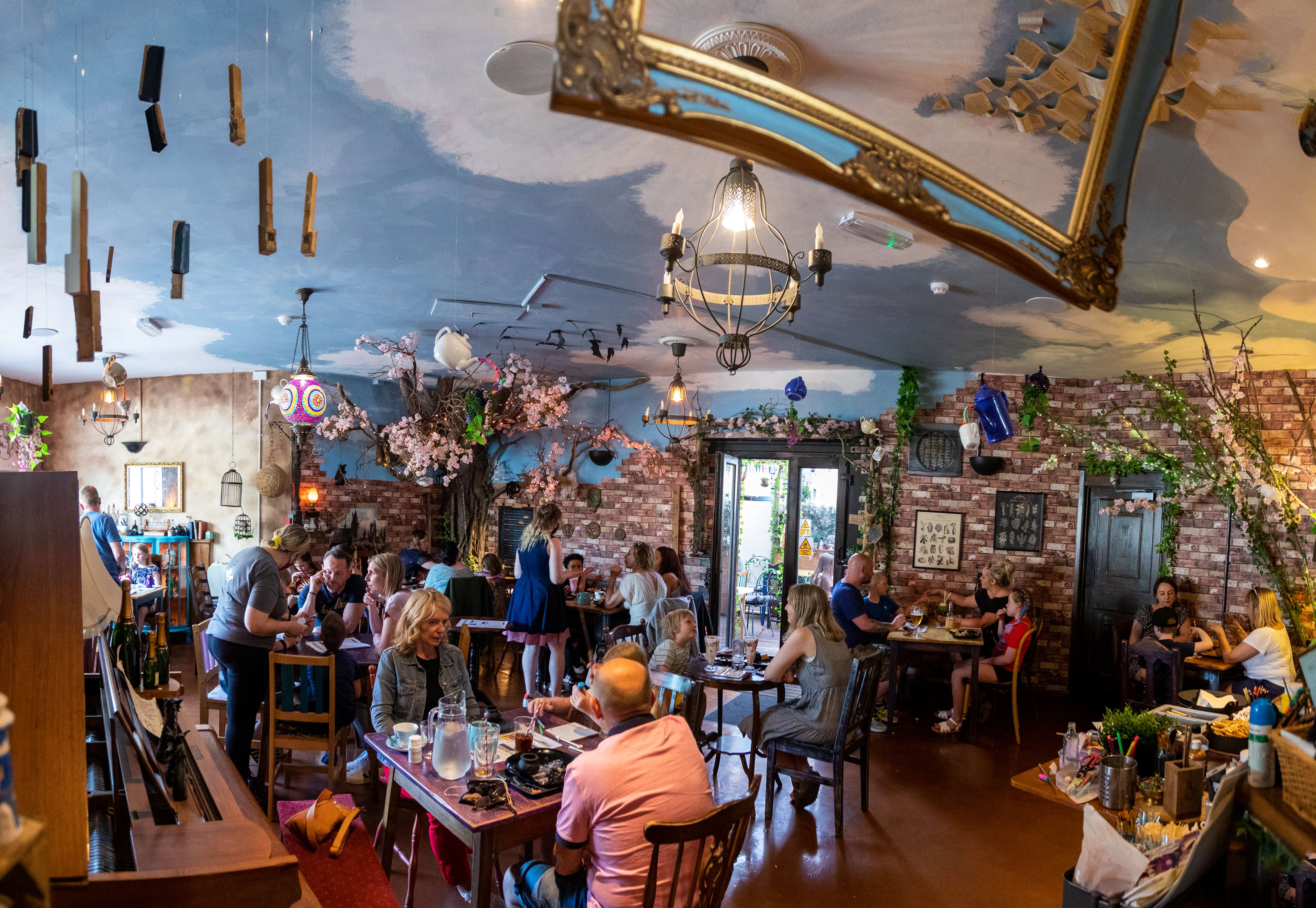 A whimsical café interior with a fantasy-inspired theme. The ceiling is painted like a blue sky with clouds and decorated with hanging objects and ornate frames. The space features exposed brick walls, artificial cherry blossom trees, and rustic wooden furniture. Several tables are occupied by people enjoying food and drinks, and the room is softly lit with decorative chandeliers and warm ambient lighting.