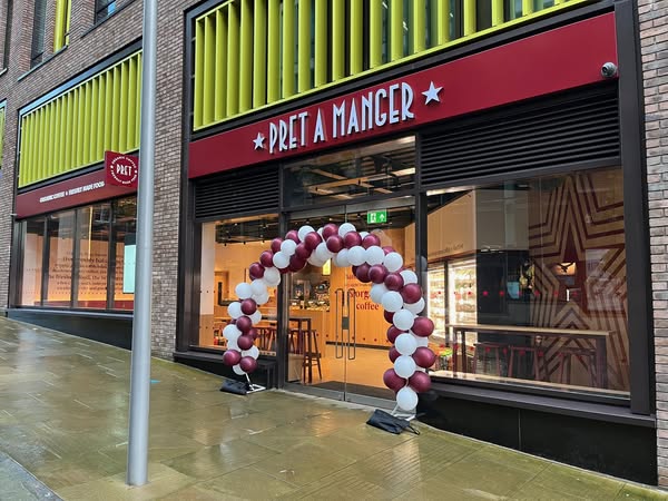 The exterior of a Pret A Manger café with a red sign displaying the name above large glass doors. A decorative arch made of maroon and white balloons stands at the entrance. The building features brick walls and bright yellow vertical panels above the storefront, and the pavement outside appears wet, suggesting recent rain.