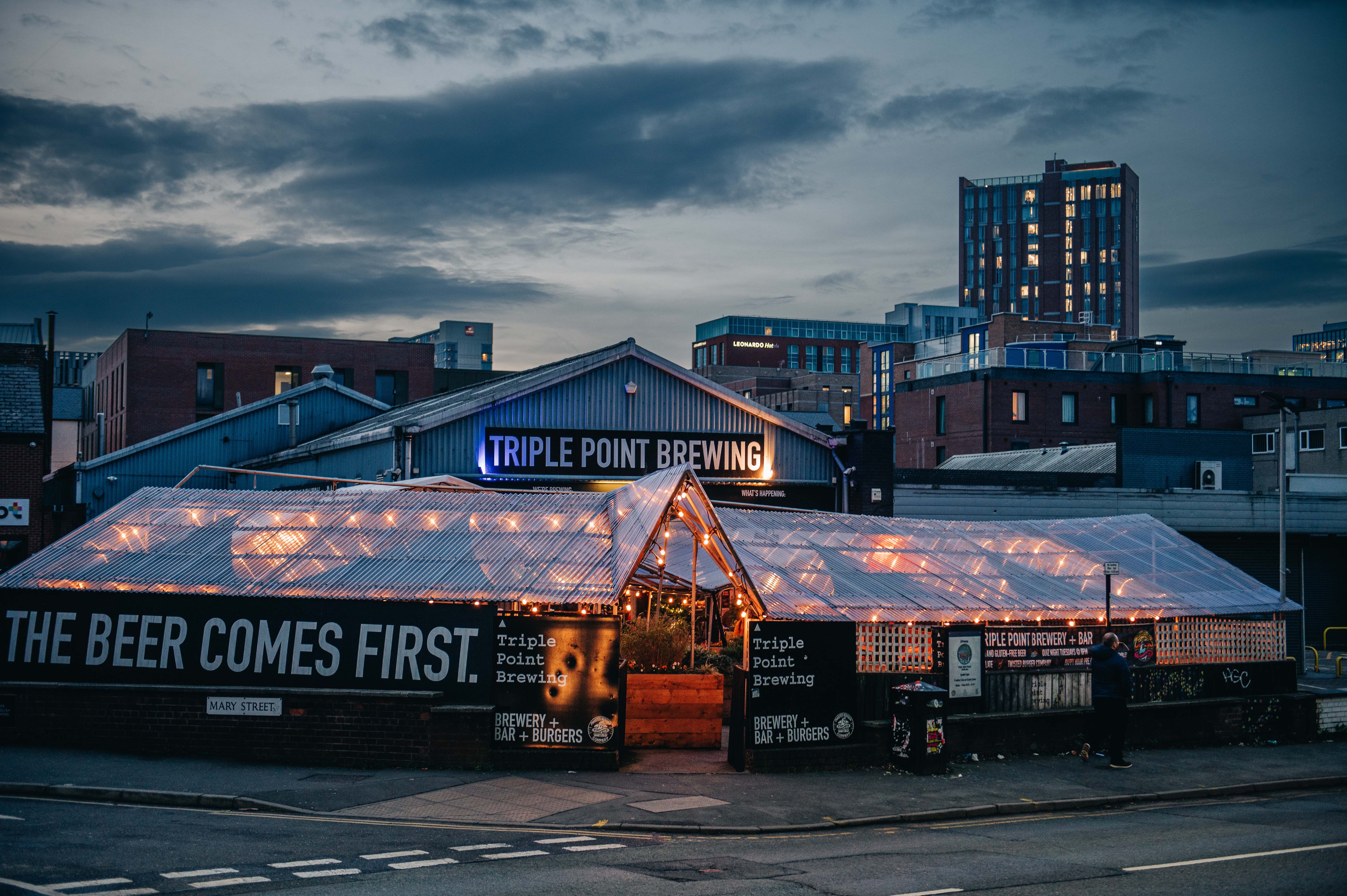 Triple Point Brewery exterior with twinkly lights at front beer garden and 'The Beer Comes First' sign. Sun is setting with cityscape in background.
