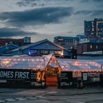 Triple Point Brewery exterior with twinkly lights at front beer garden and 'The Beer Comes First' sign. Sun is setting with cityscape in background.