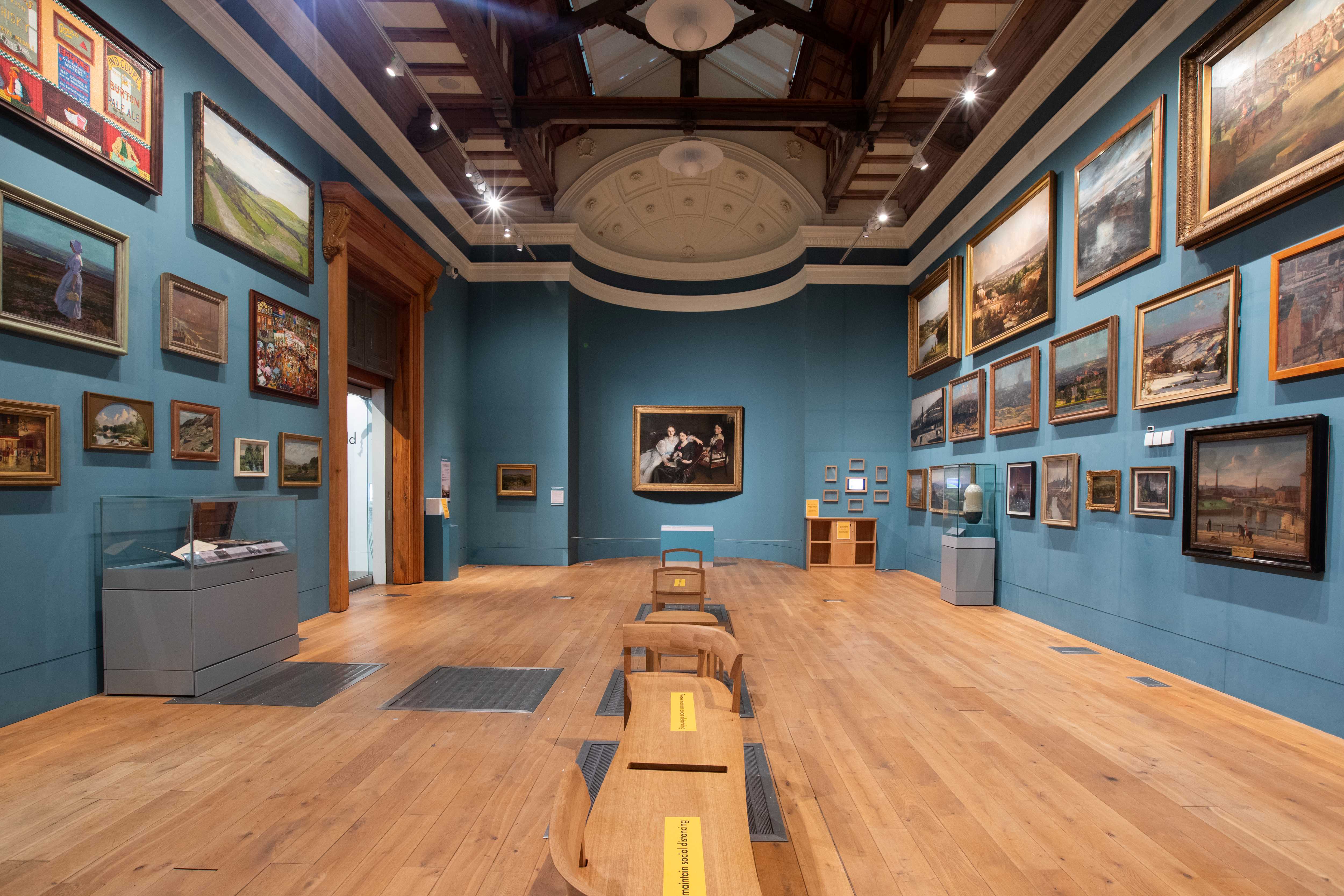 Inside one of the large galleries at the Weston Park Museum. The walls are covered in many paintings. There are also display cases against some of the walls. A row of wooden seating runs down the centre of the room.