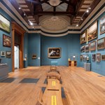 Inside one of the large galleries at the Weston Park Museum. The walls are covered in many paintings. There are also display cases against some of the walls. A row of wooden seating runs down the centre of the room.