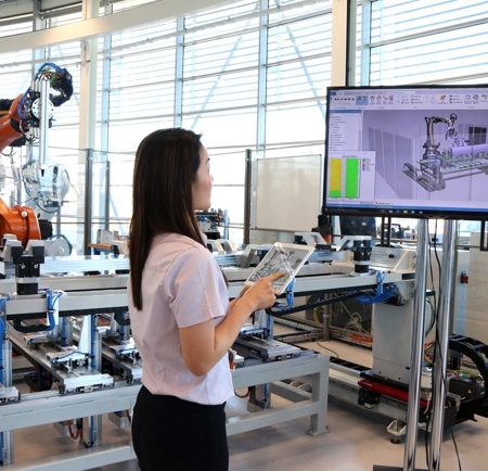 A woman is standing in front of a monitor and holding a tablet. In the background is an industrial robot arm.
