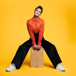 A portrait photography of Jen Brister. She is wearing a striking orange shirt, black trousers and white trainers. She is sat astride a large wooden block ans is staring directly at the camera with a neutral expression. The background is dark yellow.