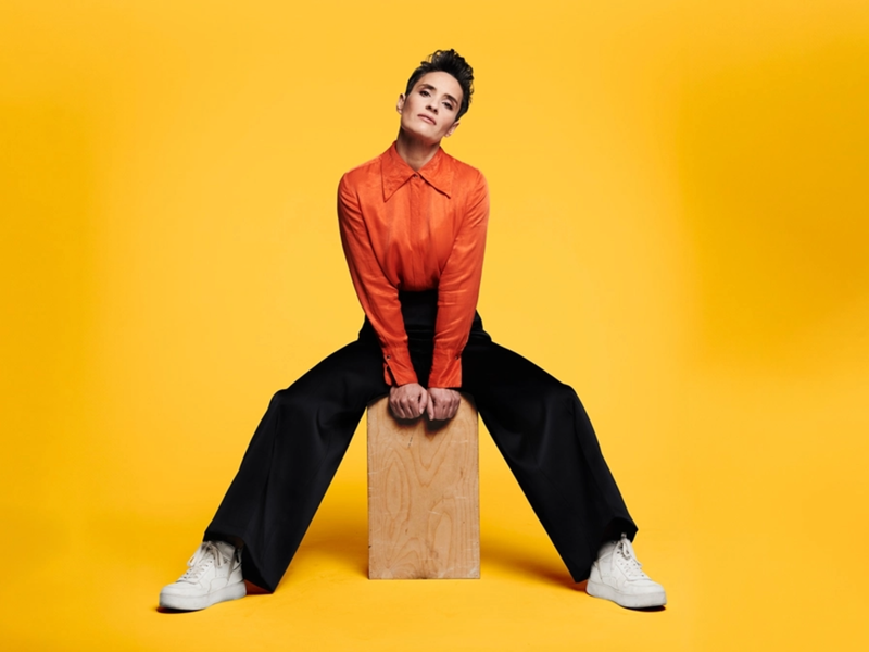 A portrait photography of Jen Brister. She is wearing a striking orange shirt, black trousers and white trainers. She is sat astride a large wooden block ans is staring directly at the camera with a neutral expression. The background is dark yellow.