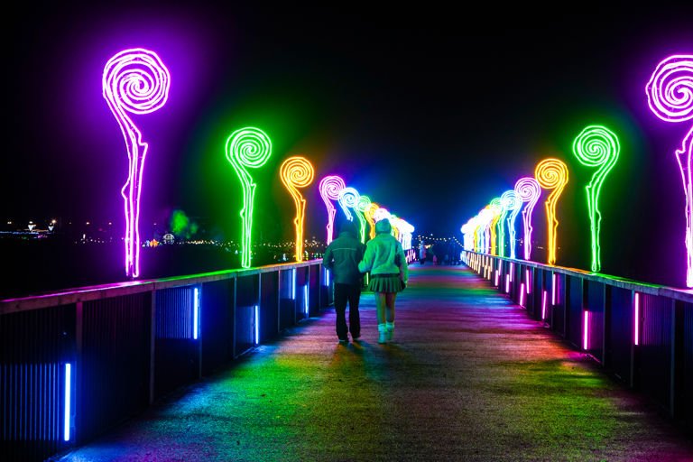 Two people walk along a brightly lit pier at night, surrounded by colorful neon light sculptures shaped like spirals. The lights glow in vibrant shades of purple, green, yellow, and pink, casting reflections on the walkway. The background is dark, with distant lights visible across the water.