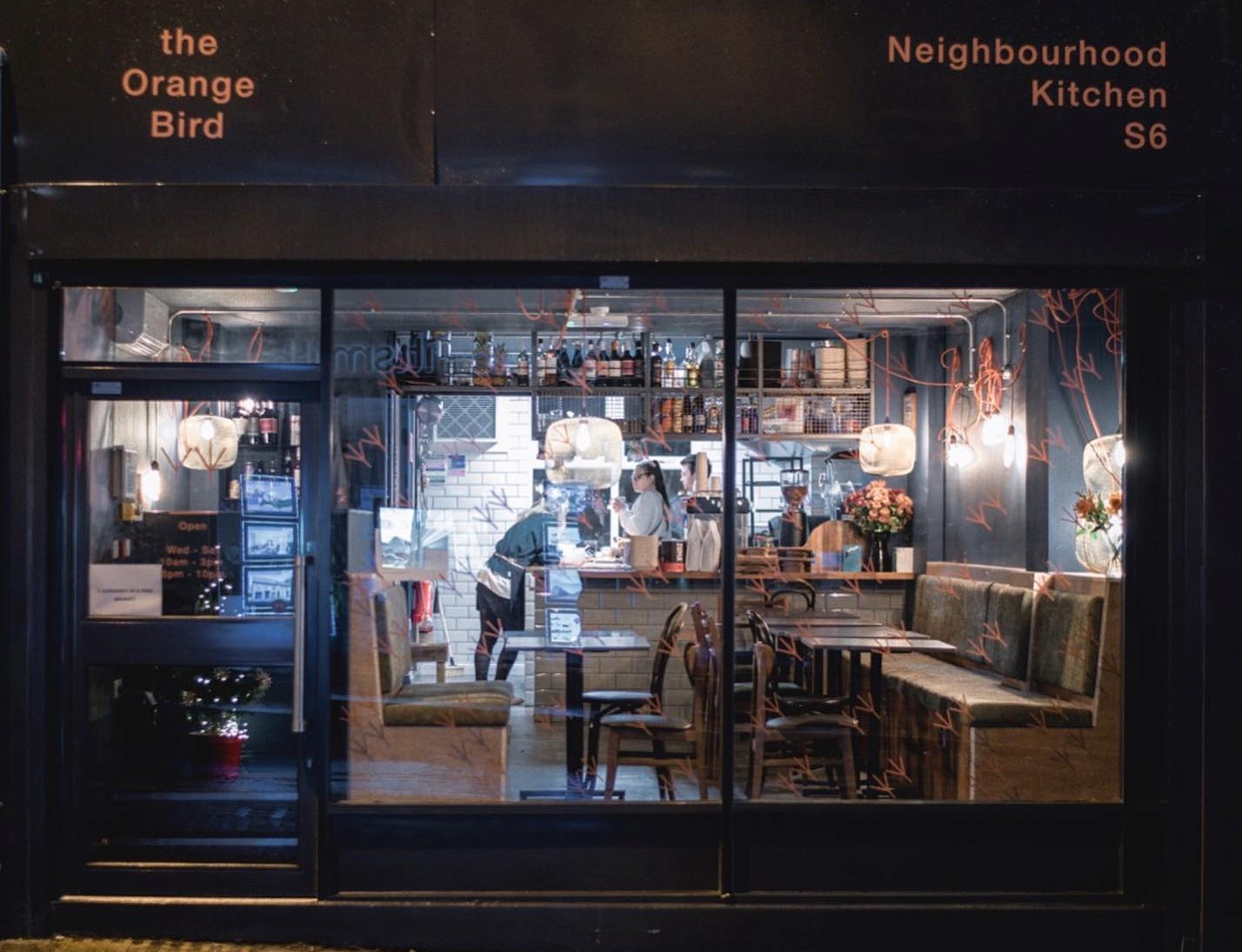Front view of The Orange Bird restaurant at night, showing large windows with a clear view of the interior dining area, pendant lights, and a bar with shelves of bottles and glassware.