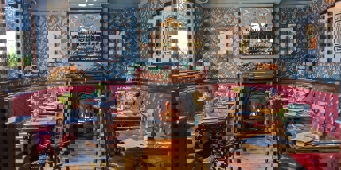  A warm, vintage‑style pub dining area with patterned wallpaper, red leather booth seating, wooden tables, and a lit fireplace beneath a decorative mirror.