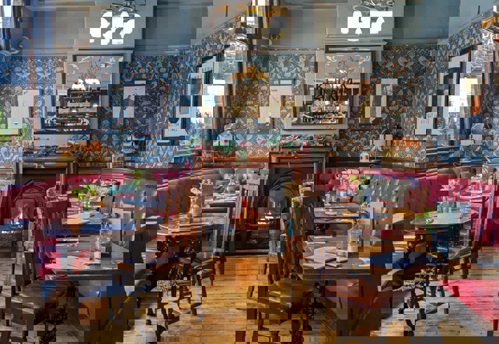  A warm, vintage‑style pub dining area with patterned wallpaper, red leather booth seating, wooden tables, and a lit fireplace beneath a decorative mirror.