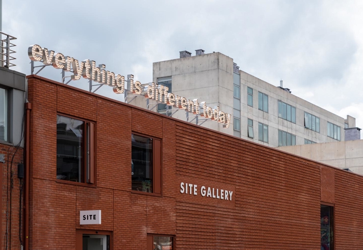 Exterior of the red brick SITE Gallery building with the gallery name displayed on the wall and an 'everything is different today' illuminated sign and cloudy sky above.