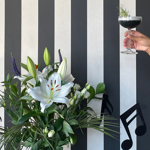 A cocktail held up over a lavish flower arrangement in front of a wall with thick black and white vertical stripes.