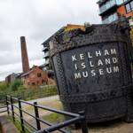 Large industrial steel vessel with rivets and the words “Kelham Island Museum” in bold letters, displayed outdoors on a paved path. Surrounding area includes railings, trees, and modern brick buildings, with a tall chimney visible in the background under a cloudy sky.