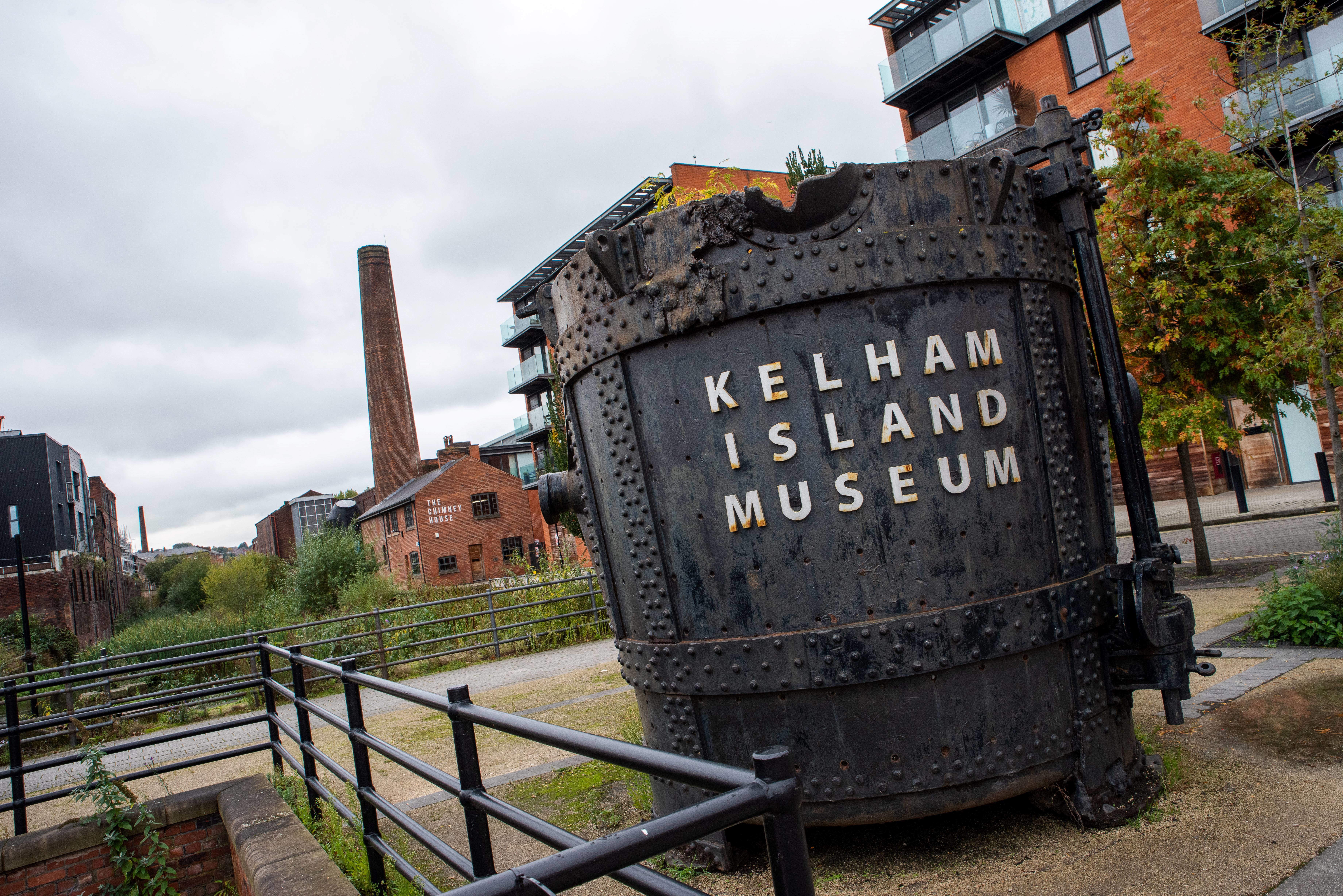 Large industrial steel vessel with rivets and the words “Kelham Island Museum” in bold letters, displayed outdoors on a paved path. Surrounding area includes railings, trees, and modern brick buildings, with a tall chimney visible in the background under a cloudy sky.