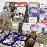 A tabletop display of stationery items such as notebooks, colouring books, illustrated prints, badges, and small accessories arranged in blue baskets and on black wire stands.