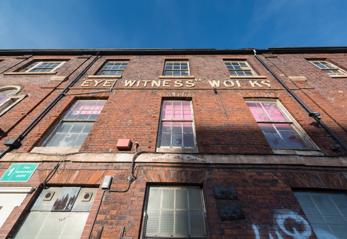 A three-story brick building with large windows, some featuring pink-tinted glass. A faded sign reads 'EYE WITNESS WORKS' across the facade. A green 'Fire assembly point' sign is visible on the left side. The sky is clear and blue.