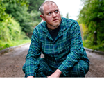 Miles Jupp, squatting down, in a country lane, wearing green checked pyjamas.