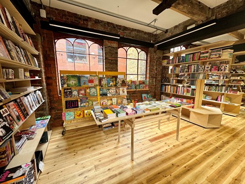 Interior of a bookstore with wooden floors and exposed brick walls featuring large arched windows. Shelves along the walls and freestanding displays are filled with colorful books, including children’s titles and graphic novels. A central table holds neatly stacked books under bright natural light.