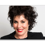  Person with short, dark, curly hair wearing a black blazer over a dark top, posed against a plain light gray background.