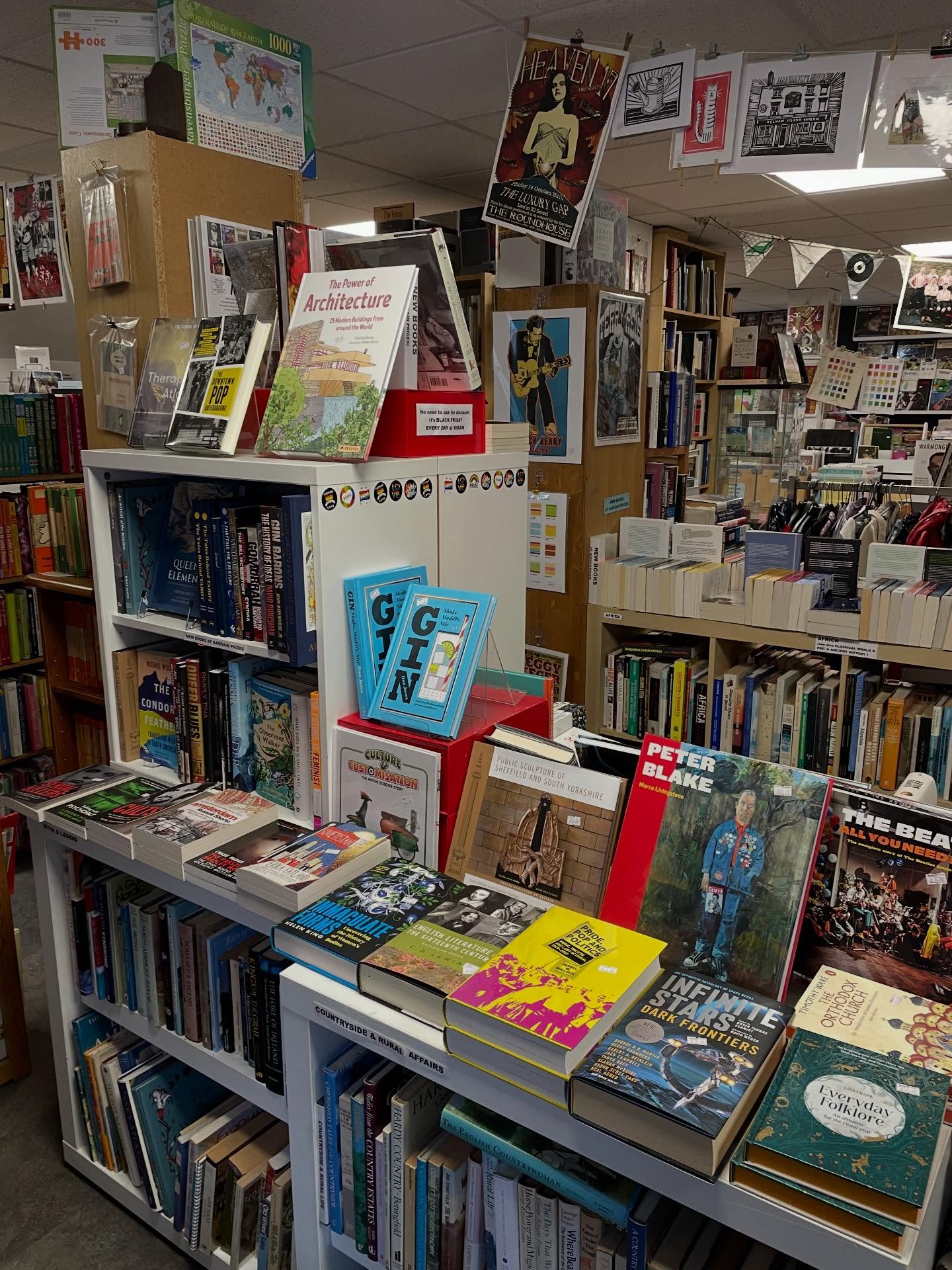 A vibrant bookshop display featuring shelves filled with books on architecture, art, and design. The foreground has colorful books stacked horizontally, including titles about gin and Peter Blake. Posters and decorative bunting hang from the ceiling, and the background shows more shelves filled with books and framed artwork.