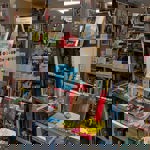 A vibrant bookshop display featuring shelves filled with books on architecture, art, and design. The foreground has colorful books stacked horizontally, including titles about gin and Peter Blake. Posters and decorative bunting hang from the ceiling, and the background shows more shelves filled with books and framed artwork.