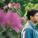 A bearded man, Kris Drever, stands amongst large plants and flowers, looking off into the distance.