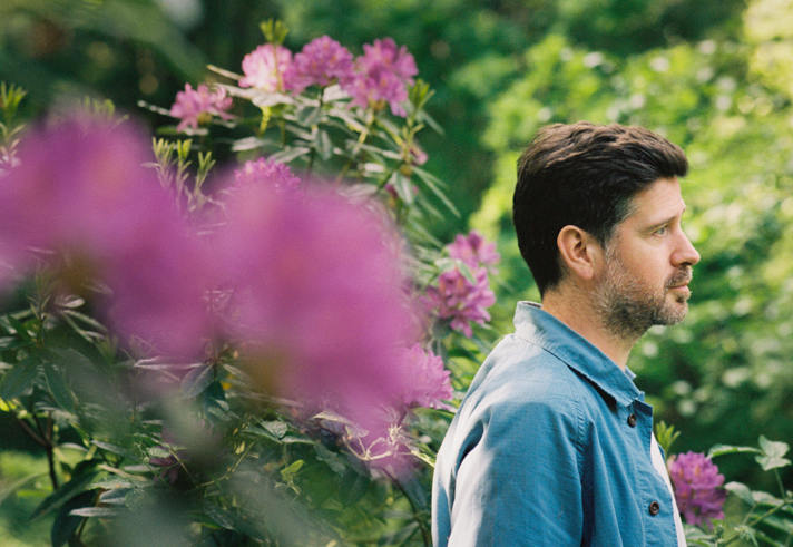 A bearded man, Kris Drever, stands amongst large plants and flowers, looking off into the distance.