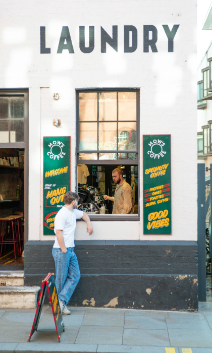 Exterior of a white-painted building with the word “LAUNDRY” in bold black letters above two windows and a doorway. The entrance is open, revealing a rustic interior with bar stools and a counter. Green signs with yellow text advertise coffee and good vibes. A person stands outside on the pavement near a sandwich board, while another is visible inside by the counter. The setting is urban, with a gated area and bicycles to the right.