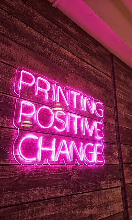 A pink neon sign that reads 'Printing Positive Change'.
