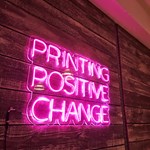 A pink neon sign that reads 'Printing Positive Change'.