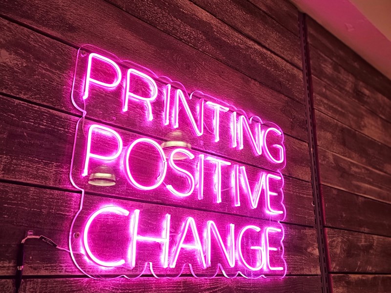 A pink neon sign that reads 'Printing Positive Change'.