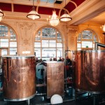 Sheffield Tap copper beer tanks. Groups of people are sat talking and drinking, with white chandeliers hanging from the ceiling, and floor to ceiling windows looking onto the station platforms.