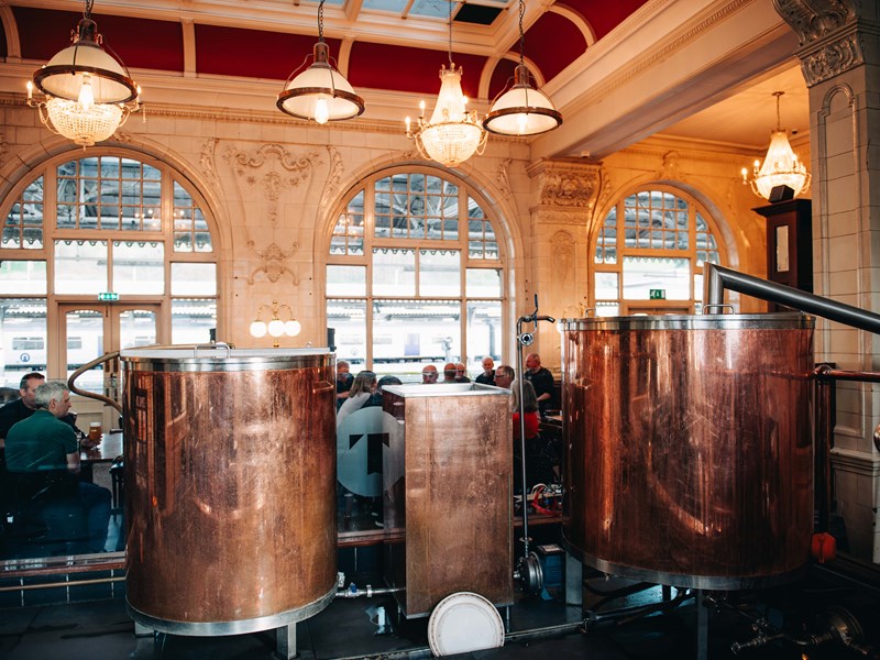 Sheffield Tap copper beer tanks. Groups of people are sat talking and drinking, with white chandeliers hanging from the ceiling, and floor to ceiling windows looking onto the station platforms.