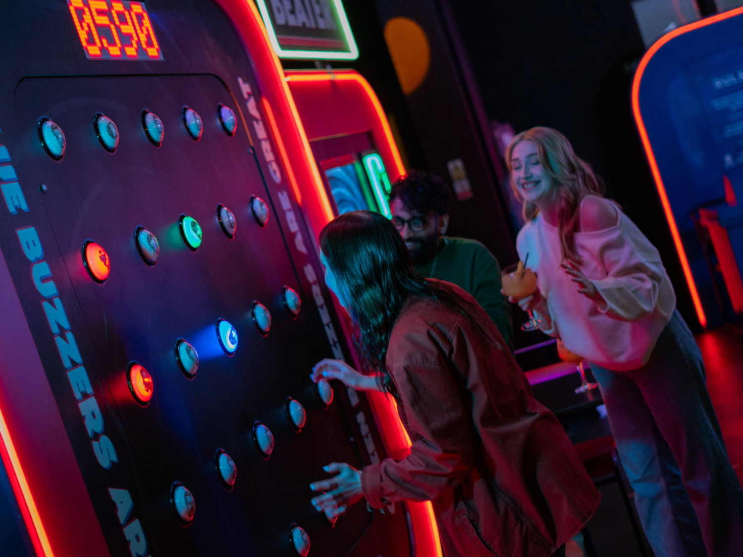 Three people at a neon-lit arcade game called “Buzzer Beater.” One person is actively playing, reaching towards illuminated buzzers on a large vertical panel displaying a score of 0590. The game features multiple round buttons glowing in blue and green, with bright signage and red lighting around the machine. Two others stand nearby, one holding a cocktail glass, watching the gameplay in a lively, colourful setting.