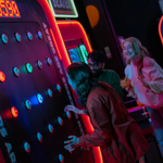 Three people at a neon-lit arcade game called “Buzzer Beater.” One person is actively playing, reaching towards illuminated buzzers on a large vertical panel displaying a score of 0590. The game features multiple round buttons glowing in blue and green, with bright signage and red lighting around the machine. Two others stand nearby, one holding a cocktail glass, watching the gameplay in a lively, colourful setting.